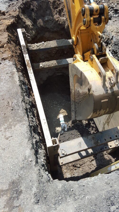 A yellow excavator digs a rectangular trench in asphalt, revealing a metal underground utility box with a pipe and valve inside—typical of sewer service in Essex County NJ. Dirt and rubble surround the excavation site.