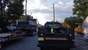 A street scene with utility trucks parked on both sides, traffic cones in the bed of one truck, and construction equipment visible—typical of sewer service in New York State. Trees and houses line the road under a partly cloudy sky.