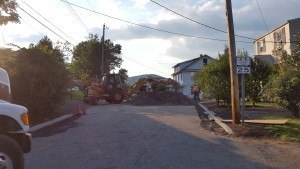 A residential street is blocked by construction equipment and piles of dirt for sewer repair in Passaic County NJ, with workers present and a speed limit sign showing 25 mph. A truck is parked on the left and trees line both sides of the road.