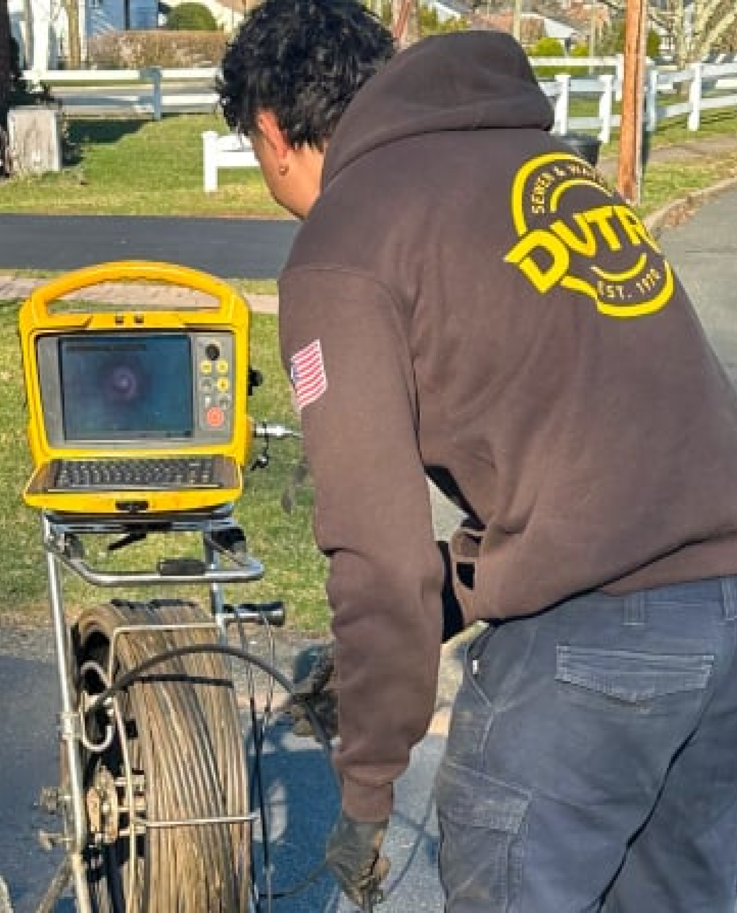 A person in a dark hoodie with "DVTA" on the back operates a pipe inspection camera for sewer repairs in Bergen County NJ, closely examining the screen that displays the camera’s internal view.