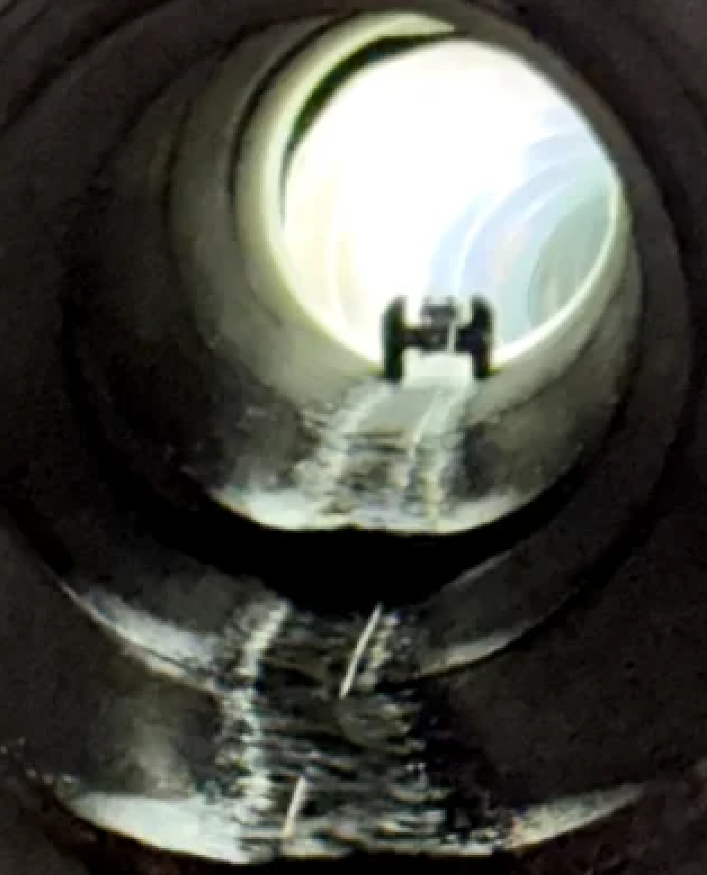View through a circular tunnel with light at the far end; a small object resembling a car or toy is silhouetted near the opening, with reflections on the wet floor—hinting at recent sewer repair in Upstate NY.