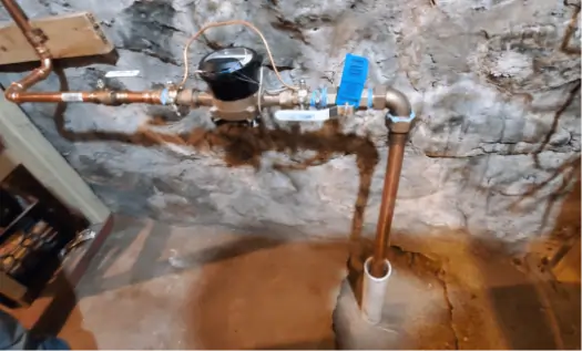 A water meter and copper plumbing pipes, part of sewer service in Essex County NJ, are installed against a rough, unfinished stone basement wall. One pipe runs vertically into a white PVC sleeve at the floor. Blue tags are attached to some of the pipes.