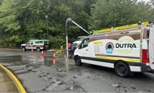 A Dutra plumbing service van and another utility truck are parked on a wet street near orange cones, as workers perform sewer service in Essex County NJ beside a wooded area.
