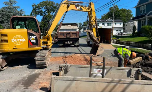 A construction worker bends over near a trench while a yellow excavator and dump truck are stationed on a residential street, showcasing sewer service in Bergen County NJ, with houses in the background.