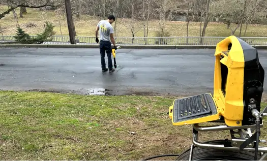 A person uses a yellow ground-penetrating radar device on a paved road, possibly for sewer repairs in Bergen County NJ, while another yellow electronic device with a keyboard sits on the grassy roadside. Trees and a fence are in the background.