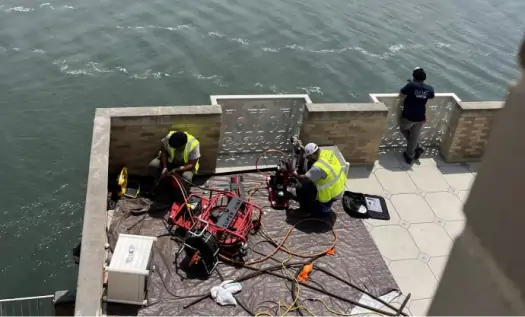 Two workers in safety vests operate equipment on a riverside terrace, possibly handling sewer repair in Upstate NY, with cables and tools spread out on a tarp. A third person stands by the railing, looking out over the water.